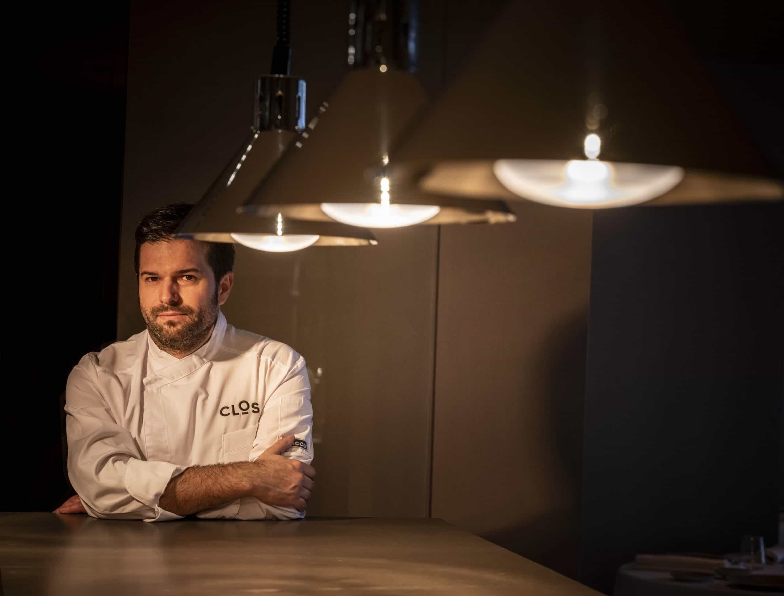 Retrato del chef Marcos Granda apoyado sobre una mesa a medida bajo iluminación cálida en su Restaurante Clos de Madrid.